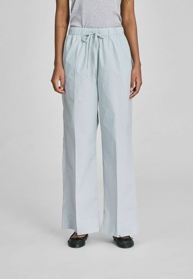 Személy világoskék, széles szárú, húzózsinóros nadrágot és fekete balerina cipőt visel, egyszerű világos háttér előtt áll.