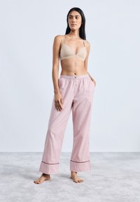 Beige háromszög alakú bralette, amely világos rózsaszín kockás pizsamával van párosítva, gombbal és piros díszítéssel az alján.