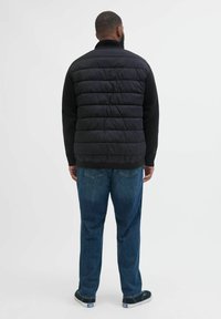 Schwarze Steppweste mit horizontalem gestepptem Design, kombiniert mit einem schwarzen langärmligen Shirt und dunkelblauen Jeans, vor einem einfarbigen Hintergrund.