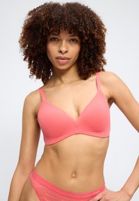 Kvinde med krøllet hår iført en koralstribet bh og matchende blonderundertøj, stående med den ene hånd på hoften.