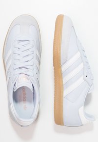 Světle modré sportovní boty s bílými akcenty, ze kůže a semiše, s kulatou špičkou a texturovanou podrážkou z gumy barvy žvýkačky. Tři bílé pruhy jsou přítomny.