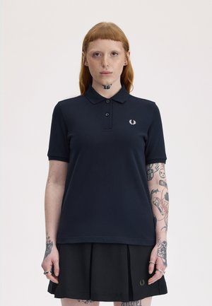 Rødhåret person med tatoveringer på nakke og arm iført en navy polo-shirt og en sort plisseret nederdel, stående foran en ensfarvet baggrund.