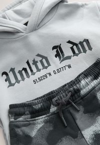 Felpa con cappuccio grigia con la scritta "Unltd Ldn" stampata in grassetto nero e coordinate sotto. Abbinata a pantaloni jogger neri con motivo mimetico. Vita elasticizzata.