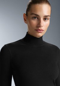 Fekete garbó felső sima textúrával, testhezálló kivitelezéssel és finom varrási részletekkel. A háttér szürke színátmenetes.