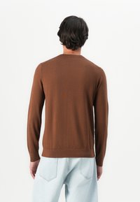 Homem com cabelo escuro, vestindo um suéter castanho de mangas longas e jeans azul claros, de pé, de frente para uma parede branca, com as mãos relaxadas aos lados.