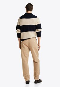 Mann in beigen Hosen, mit dunkelbraunen Schuhen und einem navyblauen und beigen gestreiften Strickpullover, steht mit dem Rücken zur Kamera.