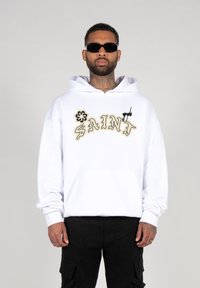 Witte hoodie met geborduurd "SAINT" in goud en zwart. Heeft een frontzak en een capuchon met trekkoord. G gedragen met zwarte cargo shorts.