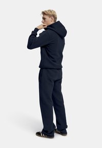 Marinblå hoodie och matchande joggers i mjukt tyg. Hoodien har en dragsko och ficka fram. Skorna är svarta med vita ränder.