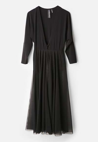 Melnā kleitā ar dziļu V veida kakla izgriezumu, garām piedurknēm un plūstošu tilla svārku. Augšējā daļa ir pieguļoša, pretstatā brīvajai apakšdaļai.