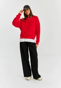 Červená mikina s textúrovaným nápisom, vrstvená na bielej košeli. Skombinovaná so voľnými čiernymi nohavicami a čiernym šiltovkou. Tenisky dopĺňajú celý outfit.