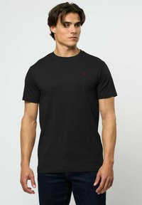 Vīrietis valkā vienkāršu melnu īsroku T-kreklu ar mazu sarkanu logotipu kreisajā krūtīs, kombinējot ar tumši zilām džinsa biksēm, stāvot pret baltu fonu.