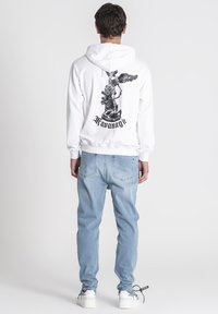 Sudadera blanca con un gráfico de ángel negro en la parte posterior, combinada con jeans de mezclilla azul claro y zapatillas blancas con acentos de marca negros.