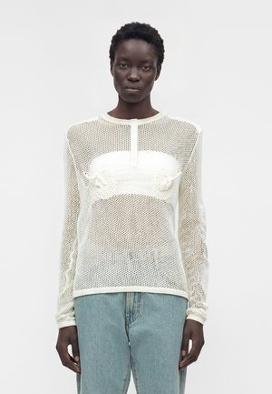 Czarna kobieta nosząca białą, długo rękawową ażurową siateczkową bluzkę z frędzlami na białym bandeau, w połączeniu z jasnoniebieskimi dżinsami, stojąca na białym tle.