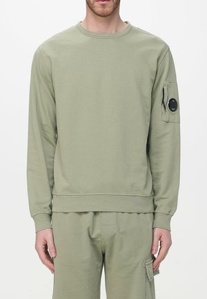 Uomo che indossa una felpa verde oliva chiaro con tasca con zip e toppa rotonda nera sulla manica sinistra, pantaloni cargo abbinati con tasca laterale.