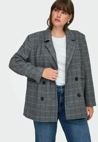 Femme portant un blazer à carreaux gris croisé sur une chemise blanche et un jeans bleu, avec des boucles d'oreilles en argent, regardant légèrement sur le côté.