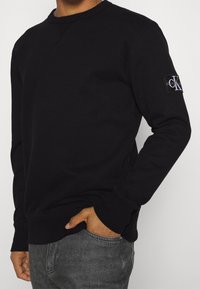 Felpa nera in cotone con collo rotondo, polsini e orlo a costine. Presenta un patch con logo sulla manica sinistra. Indossata con jeans scuri.