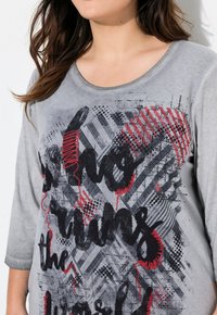 T-shirt gris à manches longues avec des motifs graphiques noirs et rouges et du texte. Matériau en mélange de coton avec un col rond et une coupe décontractée.