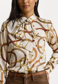 Zīda blūze ar gaišu fona krāsu, rotāta ar ķēdes un jostas rakstu zelta un zemes toņos, pabeigta ar lielu loksnes apkakli.