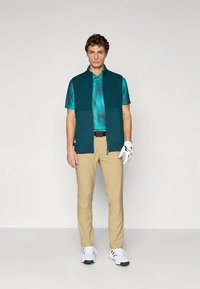 Golfas apranga, kurios pagrindinis elementas yra žalsvas liemenė dėvima ant žalsvo ir mėlyno raštuoto polo marškinėlio, kartu dėvimi smėlio spalvos kelnės ir balti golfo batai su juodais akcentais.