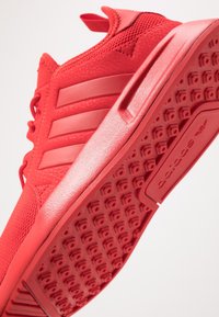 Röd Adidas-sneaker med texturerad sula, tre ränder på sidan och övre del i mesh, vinklad för att visa sid- och botten detaljer.