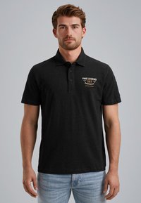 Zwart poloshirt van gladde stof, met een kraag en korte mouwen. Op de linkerborst staat gouden en witte opdruk.