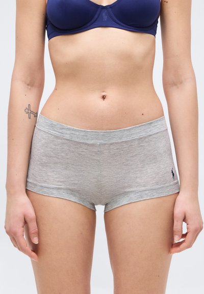 Γυναίκα που φοράει σκούρο μπλε σουτιέν και γκρι βαμβακερά boyshort εσώρουχα με μικρό λογότυπο στον αριστερό γοφό και τατουάζ σταυρού στο αριστερό αντιβράχιο.