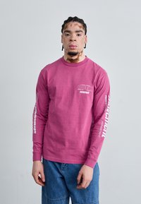 Camiseta de manga larga rosa con texto gráfico blanco en el pecho y las mangas; combinada con pantalones cortos de mezclilla; diseño sencillo, corte relajado.