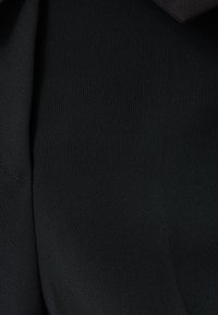Primo piano di tessuto nero, superficie liscia, con una leggera lucentezza. Presenta una cucitura e un elemento di design piegato.