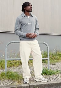 Γκρι φλις pullover με μισό φερμουάρ και μικρό λογότυπο, συνδυασμένο με άσπρα χαλαρά παντελόνια και γκρι αθλητικά παπούτσια, στέκεται δίπλα σε μια μεταλλική κουπαστή.