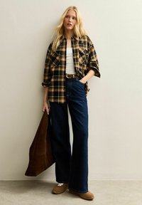 Brązowa i zielona koszula flanelowa w kratkę noszona na białym podkoszulku, w połączeniu z ciemnoniebieskimi spodniami typu wide-leg. Brązowa zamszowa torba typu tote, beżowe buty wsuwane.