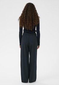 Vrouw met lang, krullend haar die een zwarte geribbelde longsleeve en wijde zwarte broek draagt, staand tegen een witte achtergrond.