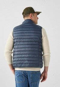 Gilet rembourré navy avec un motif matelassé horizontal, en matériau léger, col montant et design ajusté, porté sur un pull crème à côtes.