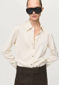 Camicia crema chiaro con bottoni e colletto, caratterizzata da bottoni dorati. Abbinata a pantaloni sartoriali marrone scuro e grandi occhiali da sole neri.