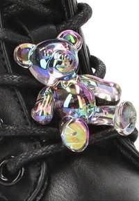 Botas negras con cordones y un charm de osito de peluche transparente y de colores del arcoíris adjunto a los cordones, que muestra formas brillantes y redondeadas en colores vibrantes.