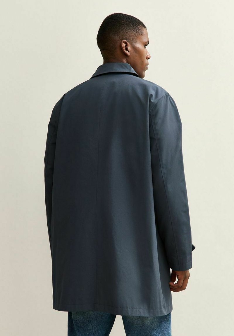 Hombre vestido con un abrigo largo gris oscuro y jeans azules, visto desde la espalda, de pie contra un fondo claro y liso.