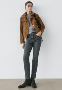 Ung kvinna som bär en brun mockajacka, grå tröja, mörkgrå jeans och svarta ankellåga stövlar, stående med ena handen i fickan mot en vit vägg.