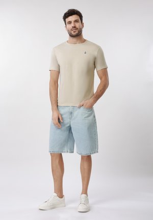Homme debout portant une chemise beige à manches courtes avec un emblème d'ancre, un short en denim bleu clair et des baskets blanches sur fond uni.