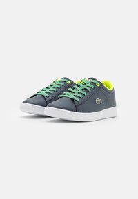 Een paar donkerblauwe sneakers met groene veters, neongele voering, witte zolen en een klein krokodillenlogo aan de zijkanten.