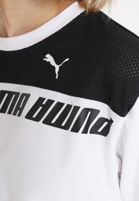 Valkoinen urheilupaita, jossa on musta verkkopaneeli olkapäissä, rohkeaa mustaa "PUMA"-tekstiä ja valkoinen PUMA-logo vasemmassa yläkulmassa.
