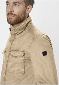 Beige jacket made of textured fabric, featuring a high collar, front pocket with flap, and a black brand patch on the shoulder.