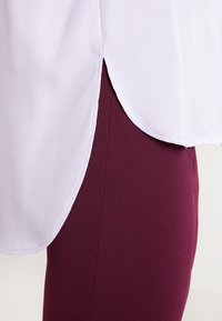 Blusa blanca y ligera con dobladillo curvado superpuesta sobre leggings color burdeos oscuro, destacando texturas suaves y capas.