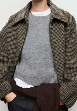 Persona que lleva una chaqueta marrón con estampado de pata de gallo sobre un suéter gris con una camisa blanca debajo, y un suéter marrón oscuro atado a la cintura.