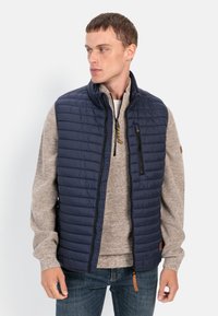 Marineblå quiltet vest med høj krave, lynlåslomme foran og brun accenttag; båret over en beige striktrøje og blå jeans.