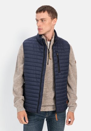 Marineblauer gesteppter Weste mit hohem Kragen, Reißverschluss-Fronttasche und braunem Akzent-Tag; getragen über einem beigen Strickpullover und blauen Jeans.