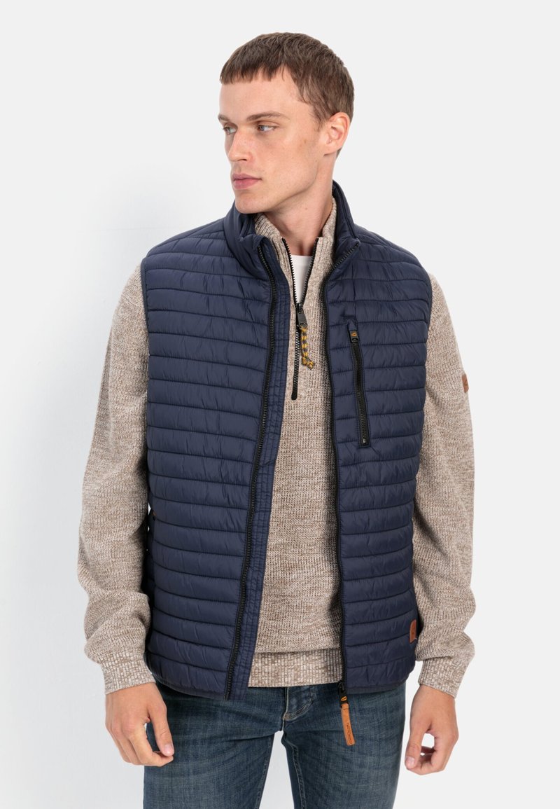 Marineblå quiltet vest med høj krave, lynlåslomme foran og brun accenttag; båret over en beige striktrøje og blå jeans.