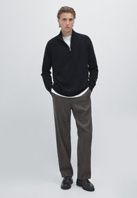 Zwart ritssluitingtrui, wit T-shirt met ronde hals eronder, grijs geblokt broek, zwarte schoenen. Effen achtergrond, staande mannelijke figuur met handen in de zakken.