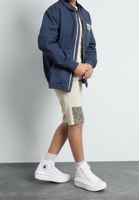 Granatowa kurtka zapinana na zamek, beżowe szorty z kratkowanym naszywką, białe wysokie trampki i szare skarpetki. Czyste linie, swobodny krój i prosty design.