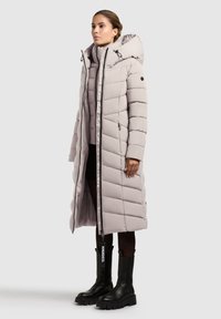 Svetlo šedý dlhý puffový kabát s kapucňou, ktorý má horizontálne prešívané vzory, predný zips a bočné vrecká. Nosený s čiernymi robustnými topánkami.