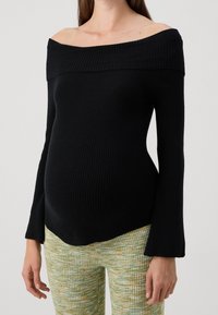 Svart off-shoulder ribbad topp med långa klockärmar, figurnära silhuett och textil med struktur, kombinerad med ljusa mönstrade byxor.