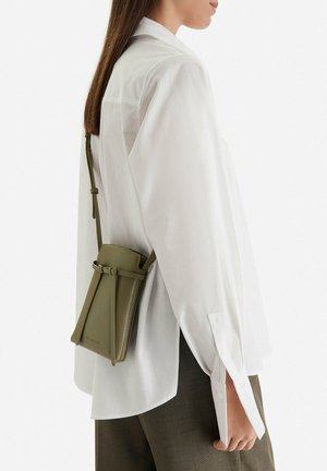 Mujer con camisa blanca y pantalones marrones llevando un pequeño bolso bandolera verde oliva con una correa delgada y ajustable.